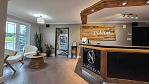 A hotel reception area with a wooden reception desk, a seating area with chairs and a round table, and a large window providing a view of the outdoors. - Hôtel Napoléon (Gatineau)