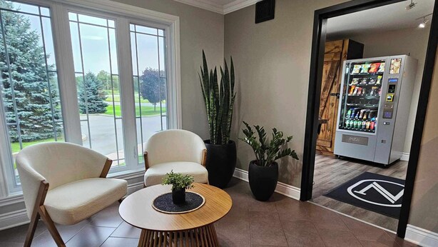 A modern lobby with a round wooden table, two white armchairs, and a potted plant. A vending machine is visible in the background. - Hôtel Napoléon (Gatineau)
