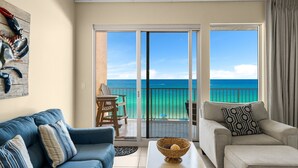 A room with a blue sofa, a beige armchair, and a dining table with a bowl. There is a sliding glass door leading to a balcony with a view of the ocean. - Beachfront resort condo w/ Gulf balcony views (Panama City Beach)