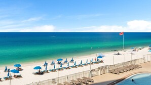 A beach with sun loungers and umbrellas, a pool, and a flag on a pole. - Beachfront resort condo w/ Gulf balcony views (Panama City Beach)