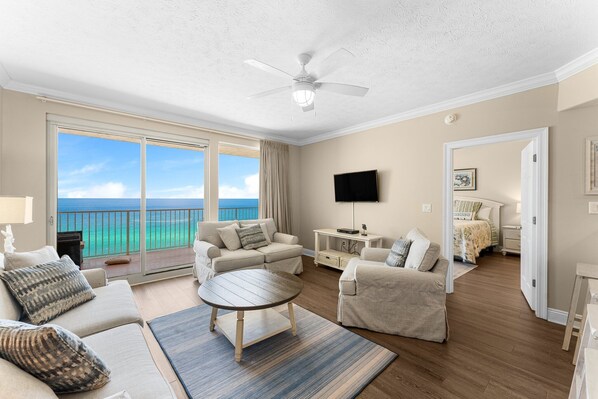 A spacious living room with a large sliding glass door leading to a balcony with a view of the ocean. There is a ceiling fan, a flat-screen TV, and a cozy seating area with a coffee table.