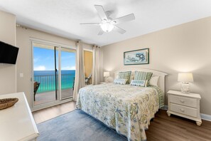 A bedroom with a bed, a nightstand, a ceiling fan, a TV, and a balcony with a view of the ocean.