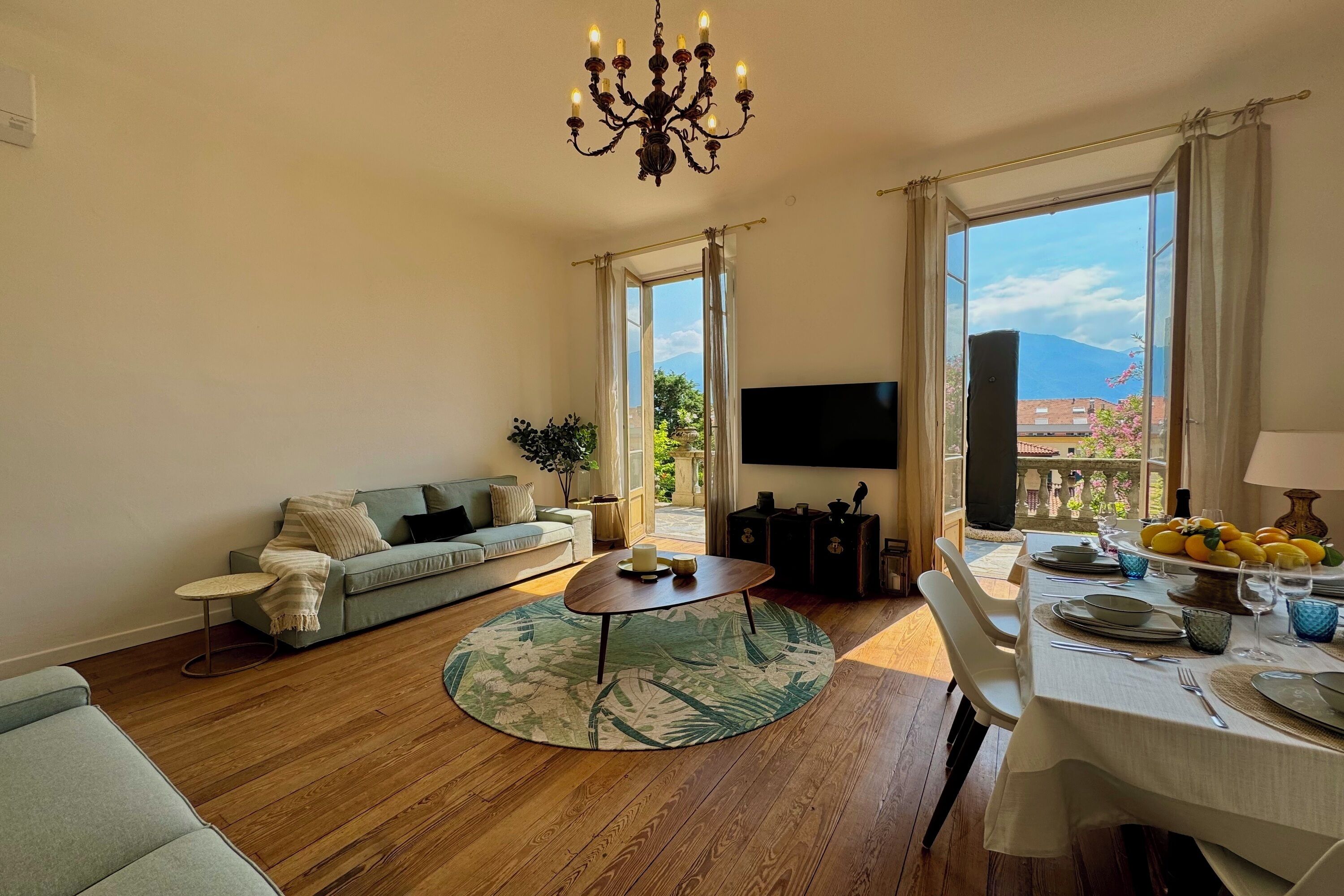 A living room with a sofa, coffee table, and a TV. There is a balcony with a table set for dining.