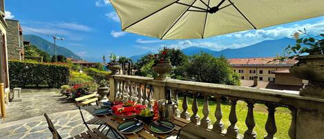 Una terraza con suelo de piedra, bancos de madera y una mesa dispuesta con platos, cuencos y botellas. Un paraguas grande ofrece sombra.