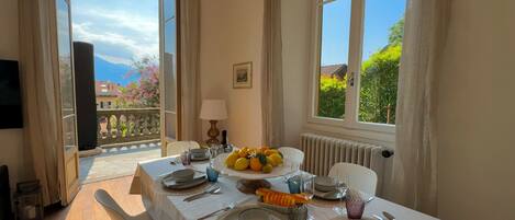A dining area with a table set for four, a fruit bowl, and a view of a balcony with a mountain in the background.