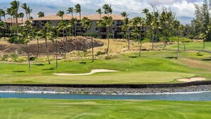 Campo de golf con un elemento acuático, palmeras y un edificio con múltiples balcones al fondo.