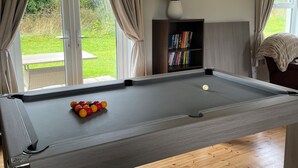 A pool table with a pool cue and balls, a bookshelf with books, and a view of the outdoors through a window.