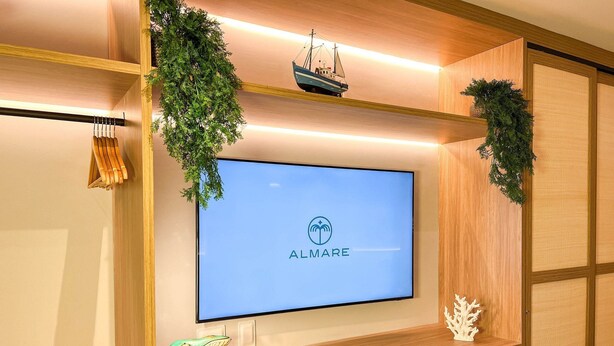 A modern hotel room with a large flat-screen TV mounted on a wooden wall, a small ship model, and decorative greenery. - Setai Yacht Luxxor - By Almare Flats (João Pessoa)