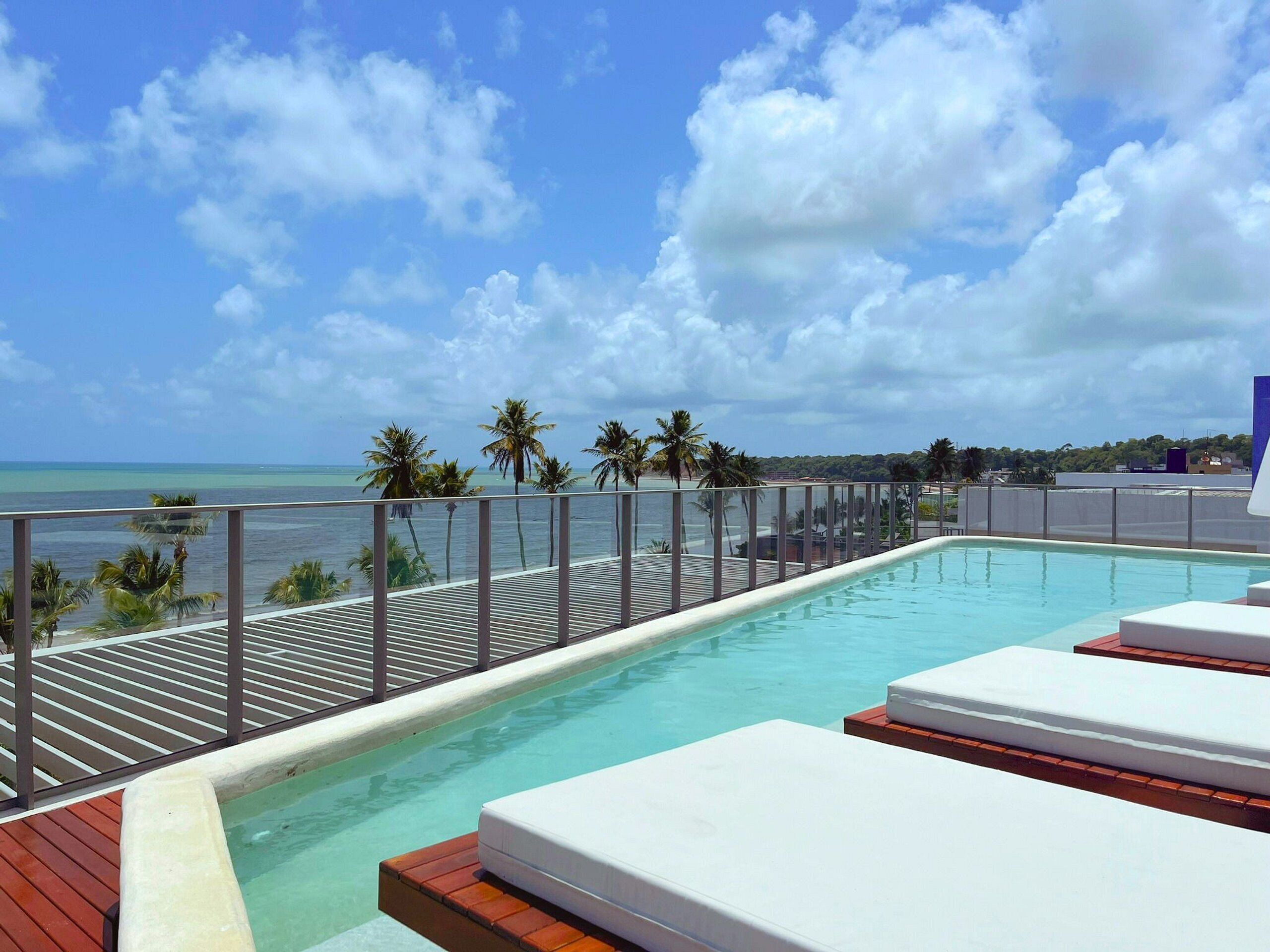 Piscina infinita no terraço com espreguiçadeiras, palmeiras e vista para o oceano.