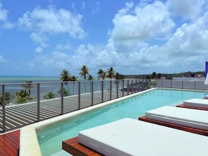 A rooftop infinity pool with lounge chairs, palm trees, and a view of the ocean.