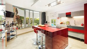 A modern kitchen with a central island, a dining area with bar stools, a built-in TV, and a large window offering a view of the outdoors. - Villa Tiberio 6+2, Emma Villas (Genzano di Roma)