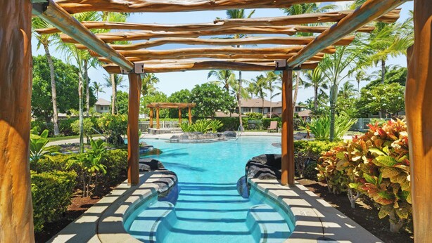Uma área de piscina com pérgola de madeira, cercada por plantas tropicais e palmeiras.