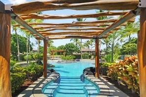 A pool area with a wooden pergola overhead, surrounded by tropical plants and palm trees.