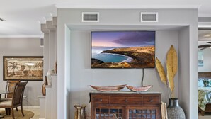 Uma sala de estar com televisão de tela plana na parede, um armário de madeira abaixo e um espaço de jantar com cadeiras e mesa.