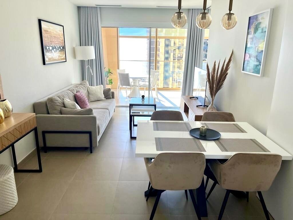 Un salon moderne avec un canapé, une table à manger et des chaises. De grandes fenêtres sont ornées de rideaux, un tableau est accroché au mur et une lampe est posée sur une table.