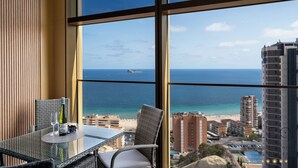 Ein Balkon mit einem Glastisch, zwei Stühlen, einer Weinflasche und zwei Gläsern. Der Blick umfasst einen Strand und eine Stadtansicht.