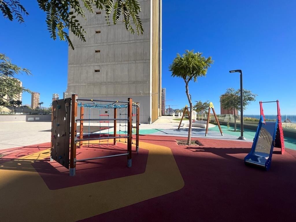 Une aire de jeux avec des structures d’escalade et une balançoire, entourée d’une zone pavée et d’arbres.
