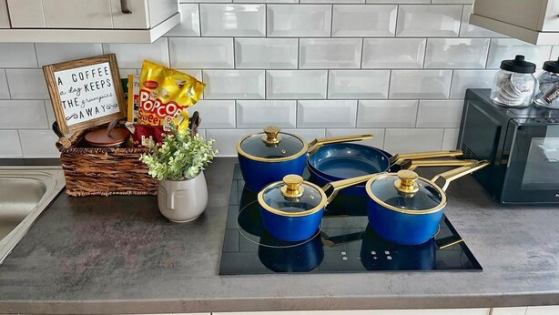 Une cuisine avec un dosseret de carreaux blancs, une cuisinière avec des casseroles bleues, un panier rempli d’en-cas et un panneau.