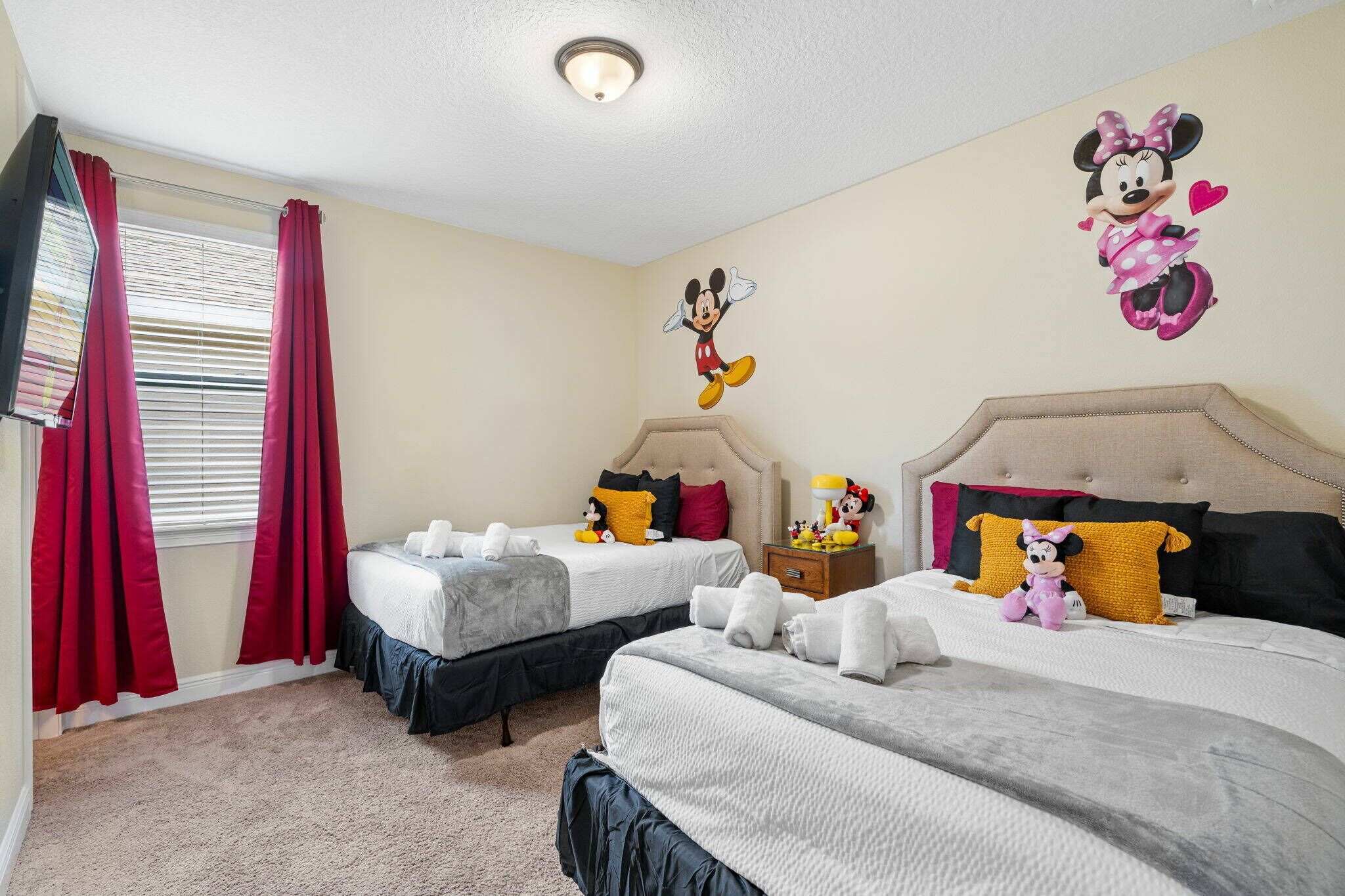 Habitación de hotel con dos camas, cortinas rojas, un adhesivo de pared de Minnie Mouse y una mesita de noche de Mickey Mouse.