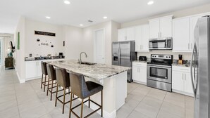 Une cuisine moderne avec des armoires blanches, des appareils en acier inoxydable, un îlot en marbre avec des tabourets, et un panneau décoratif fixé au mur.