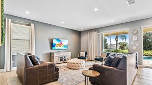 A spacious living room with a flat-screen TV, leather sofas, a coffee table, and a large window with a view of a swimming pool. - Games Room/Private Pool and Hot Tub/Beautifully furnished/Movie Room/Free Wifi (Kissimmee)