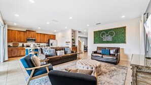 A spacious living area with a sofa, ottoman, and chairs, a kitchen with wooden cabinets, and a green wall with a logo. - Games Room/Private Pool and Hot Tub/Beautifully furnished/Movie Room/Free Wifi (Kissimmee)