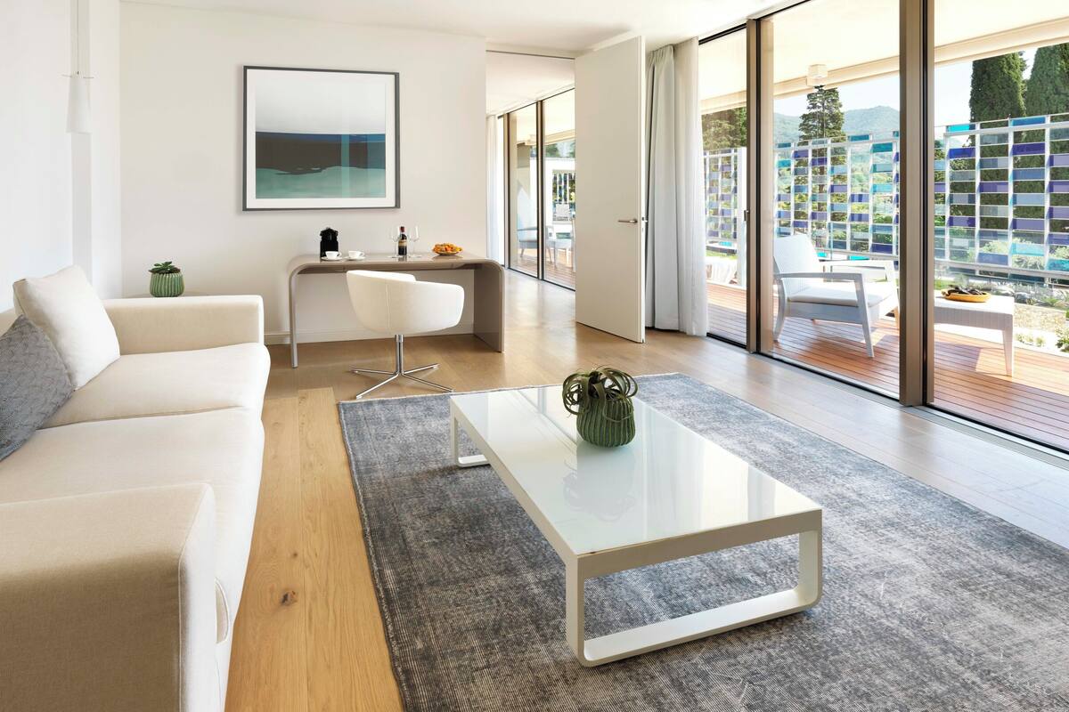 Un salon moderne avec un canapé, une table basse et un bureau. Une grande fenêtre offre une vue sur un balcon et des immeubles.