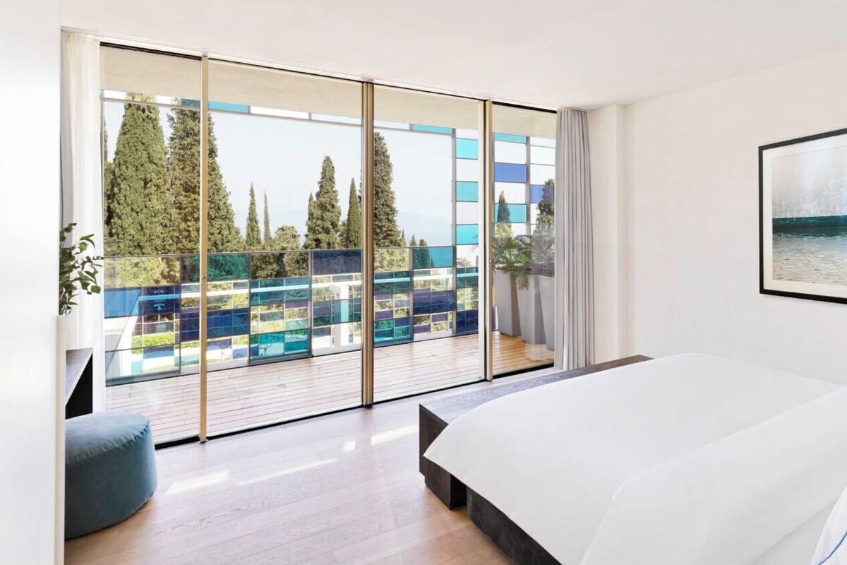 Une chambre moderne avec un grand lit, une vue sur l’extérieur grâce à de grandes portes vitrées du sol au plafond, et un tableau encadré au mur.