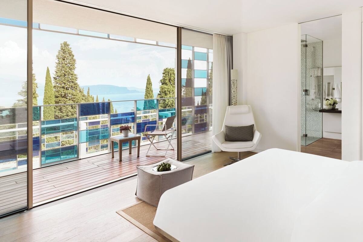 Une chambre d’hôtel moderne dotée d’un grand lit, d’un coin salon avec un fauteuil et un pouf, d’une douche en verre et d’un balcon offrant une vue sur les arbres et la mer.