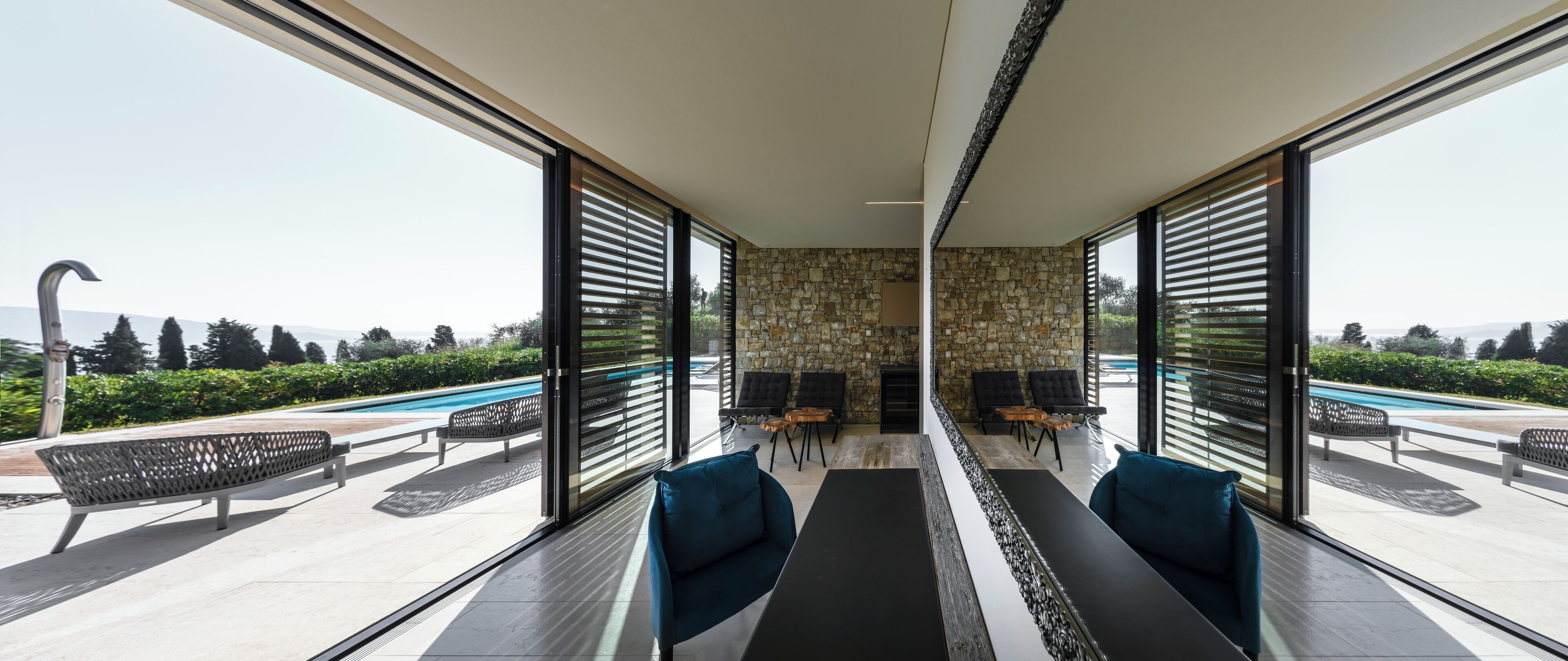 Un espace de détente moderne avec un mur en pierre, de grandes fenêtres et des sièges extérieurs situés près d’une piscine.