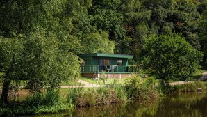 Una cabana petita i verda al costat d'un llac, envoltada per arbres frondosos.