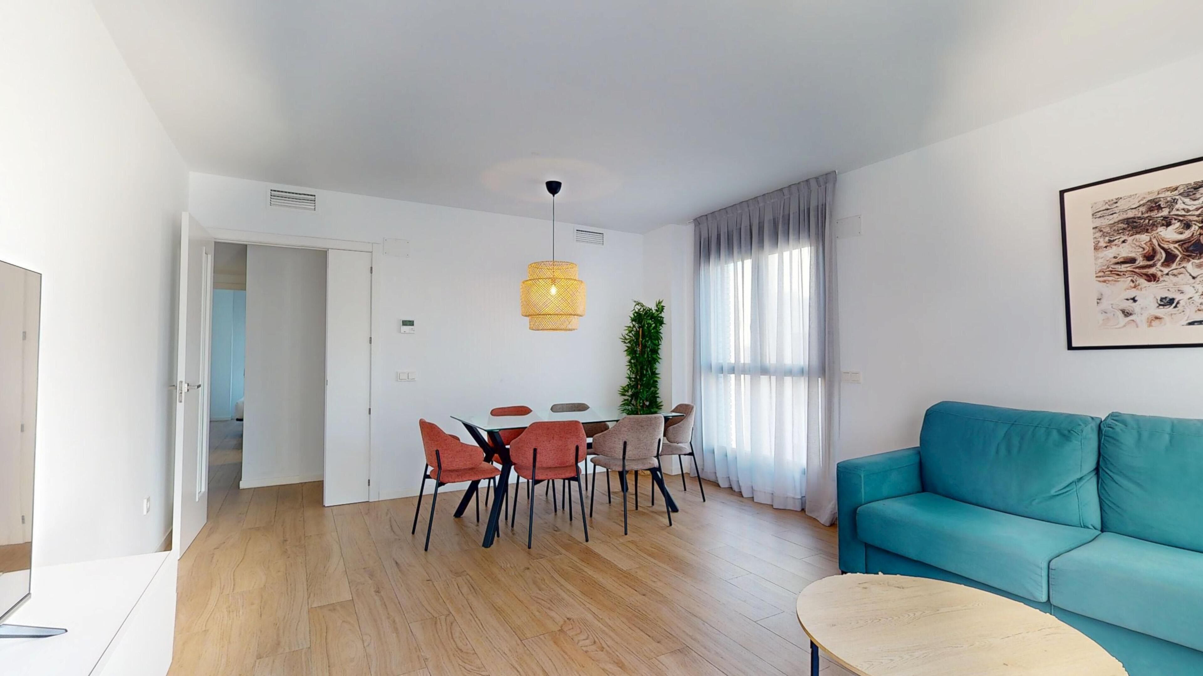 Ein modernes Wohnzimmer mit einem petrolfarbenen Sofa, einem Essbereich mit Tisch und Stühlen und einem großen Fenster mit Vorhängen.