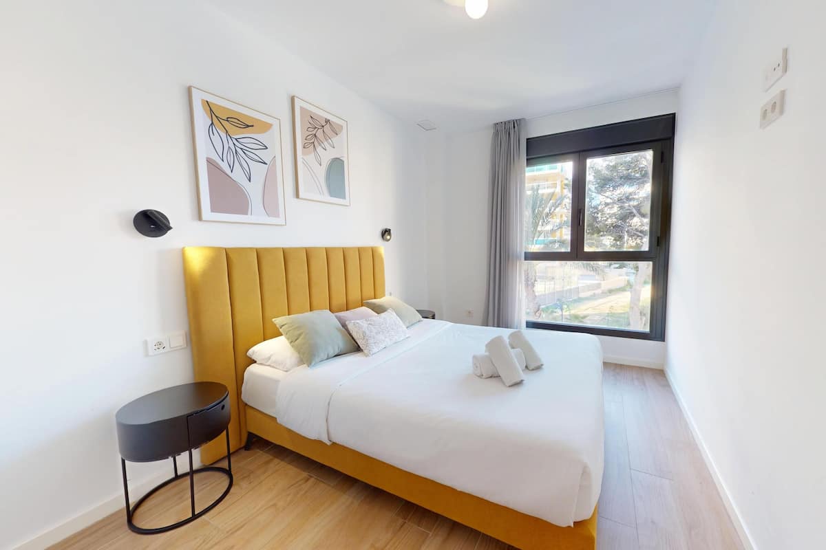 A modern hotel room with a bed, a bedside table, a window with a view, and two framed artworks on the wall.