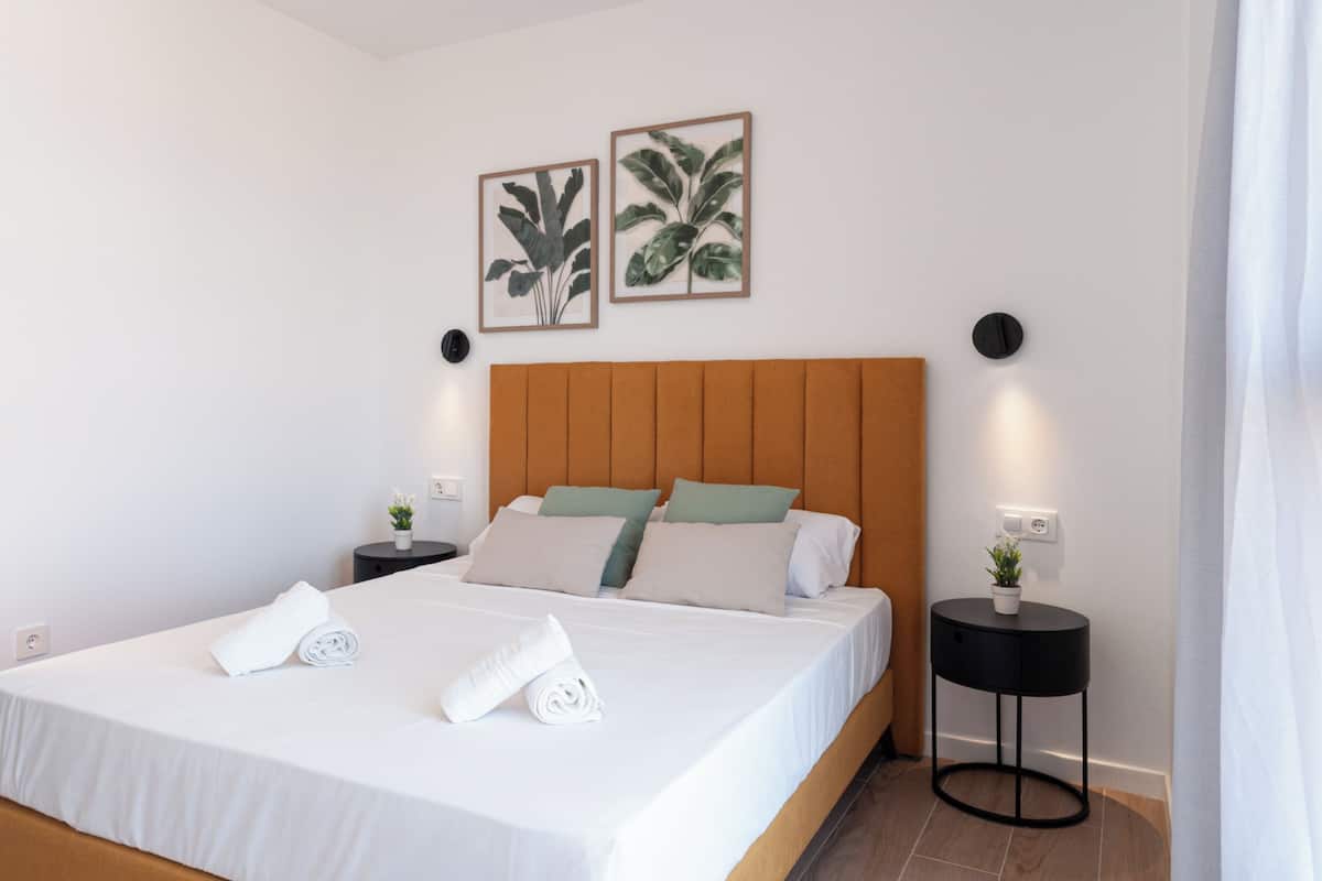 A neatly made bed with white linens and pillows, a wooden headboard, two framed plant pictures on the wall, and a small potted plant on a side table.