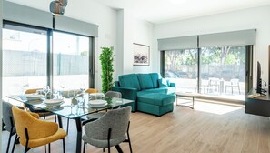 A modern living room with a glass dining table, teal sofa, and a large window offering a view of the outdoors. - Myflats Campello Beach (El Campello)