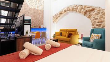 Una sala de estar moderna con un sofá amarillo, un sillón azul verdoso y una alfombra roja. Hay una zona de cocina con una pared de ladrillo y una encimera azul.