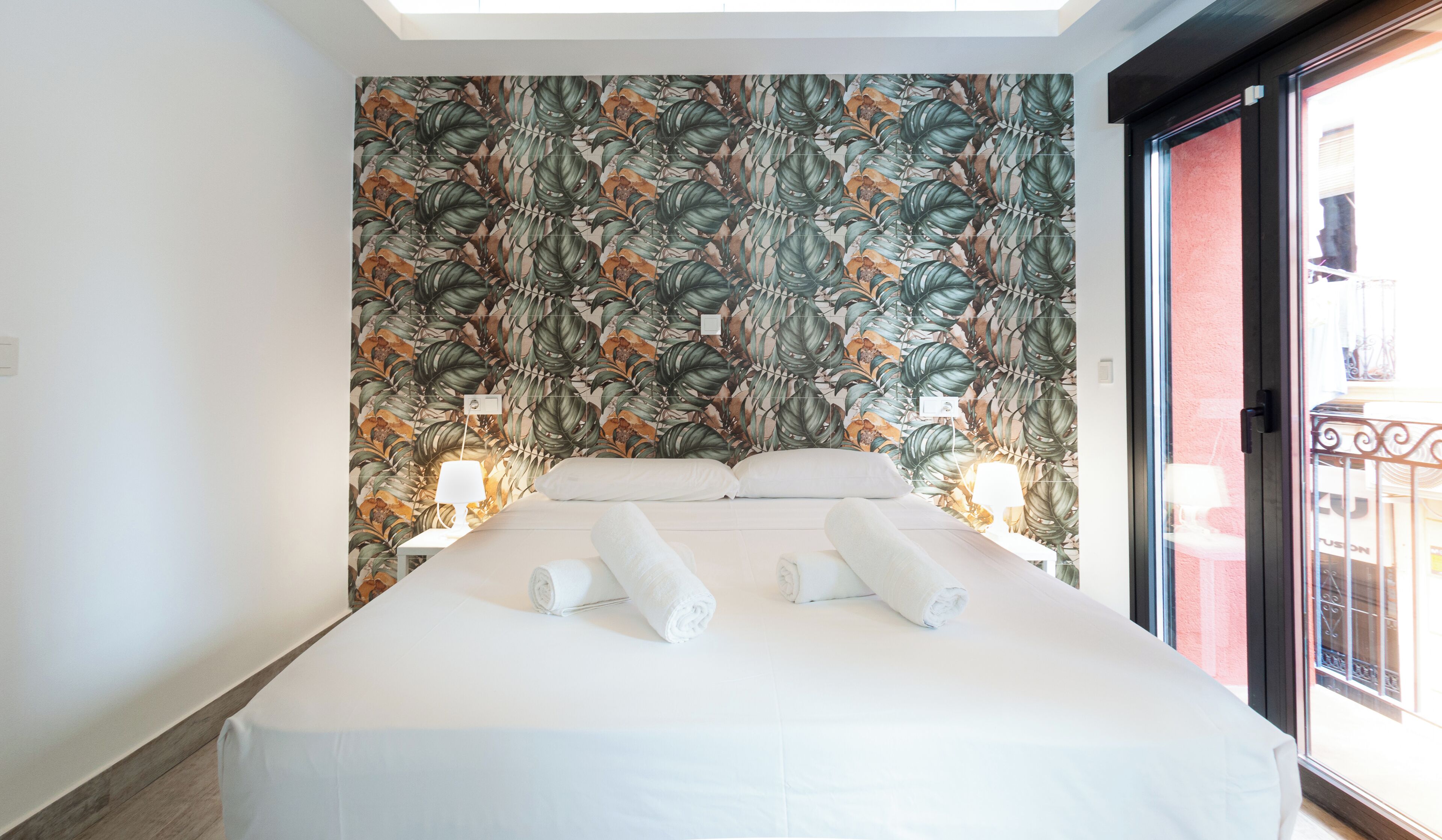 A hotel room with a large bed, two bedside lamps, and a tropical leaf-patterned wallpaper.