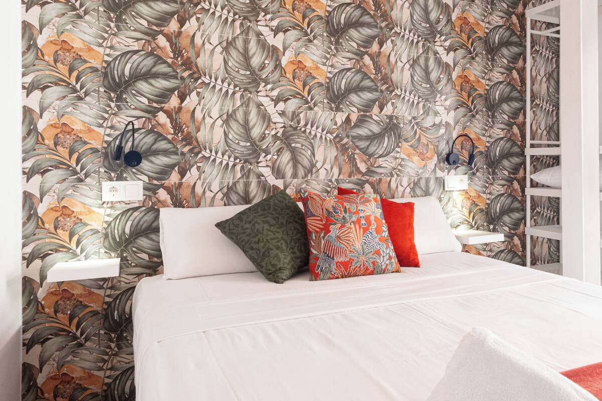 A neatly made bed with white bedding and multiple pillows, a wall with a tropical leaf pattern, and a white shelving unit to the right.