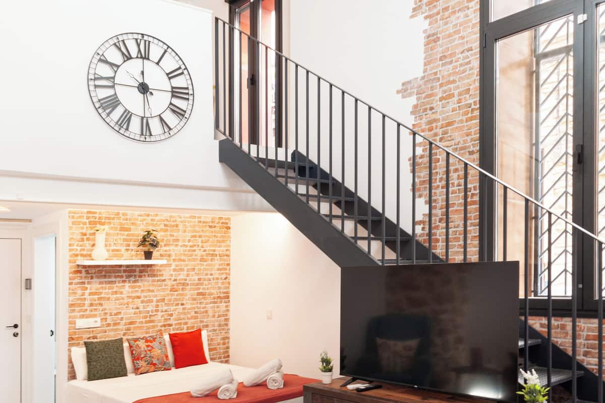 A modern loft bedroom with a brick wall, a large bed, a TV, and a staircase.