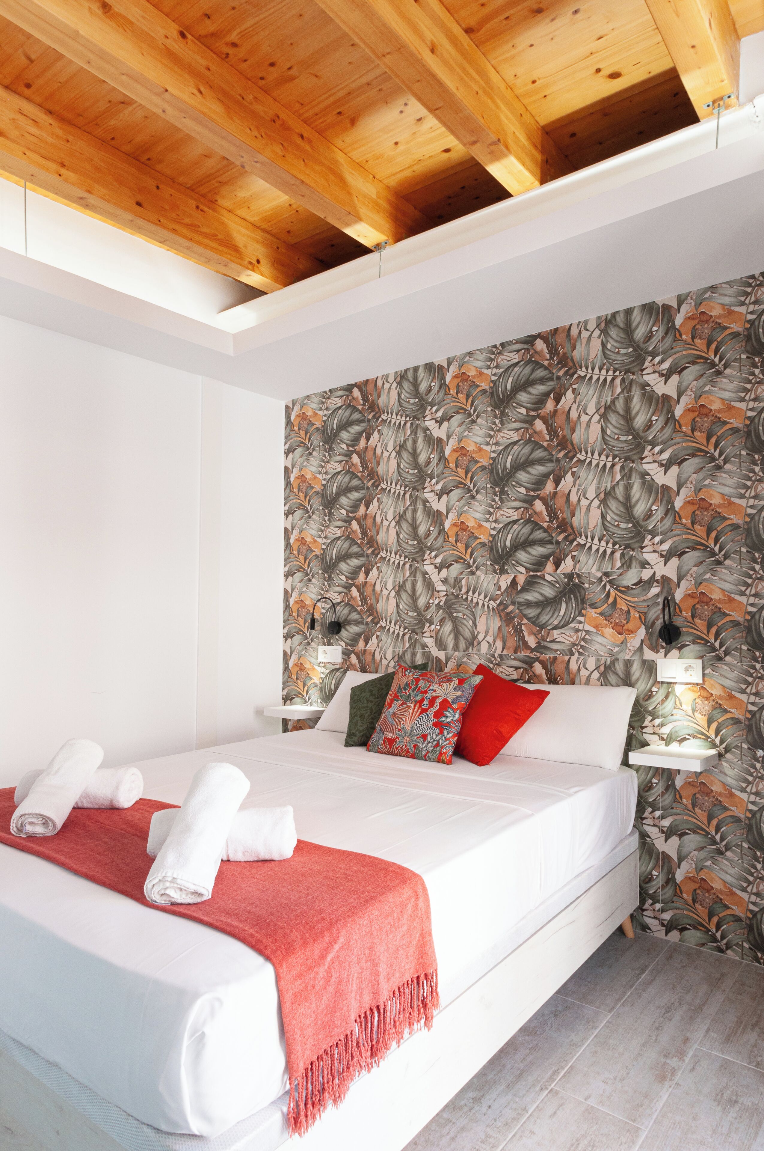 Un dormitorio con techo de madera, cama con ropa blanca, manta roja y papel tapiz floral.
