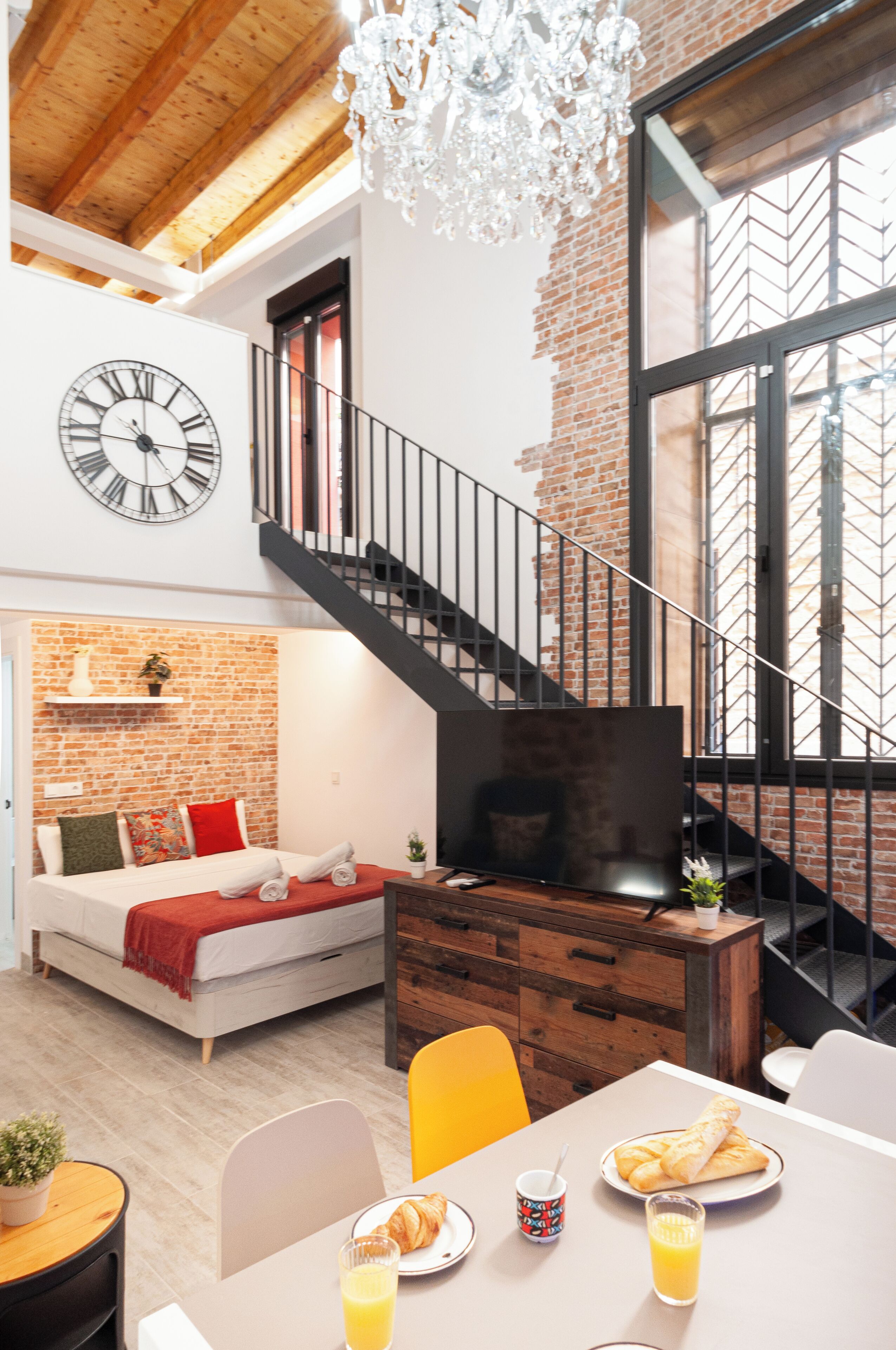 Un loft moderne avec des murs en briques apparentes, un plafond en bois, un escalier, une grande horloge, un coin salon confortable et un espace repas avec des articles pour le petit-déjeuner.