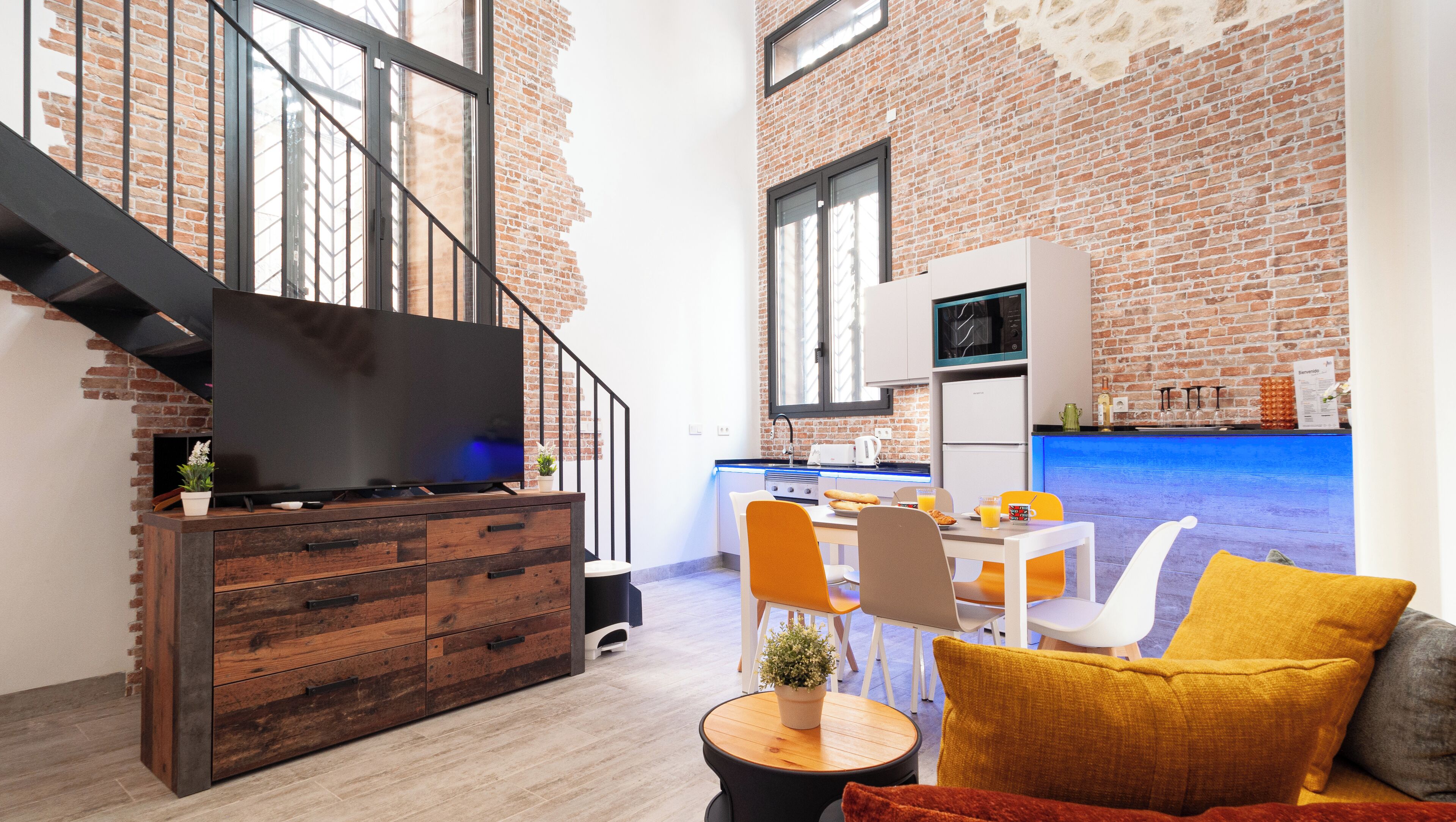 A modern kitchen with a brick wall, a flat-screen TV, and a dining area with a table and chairs.