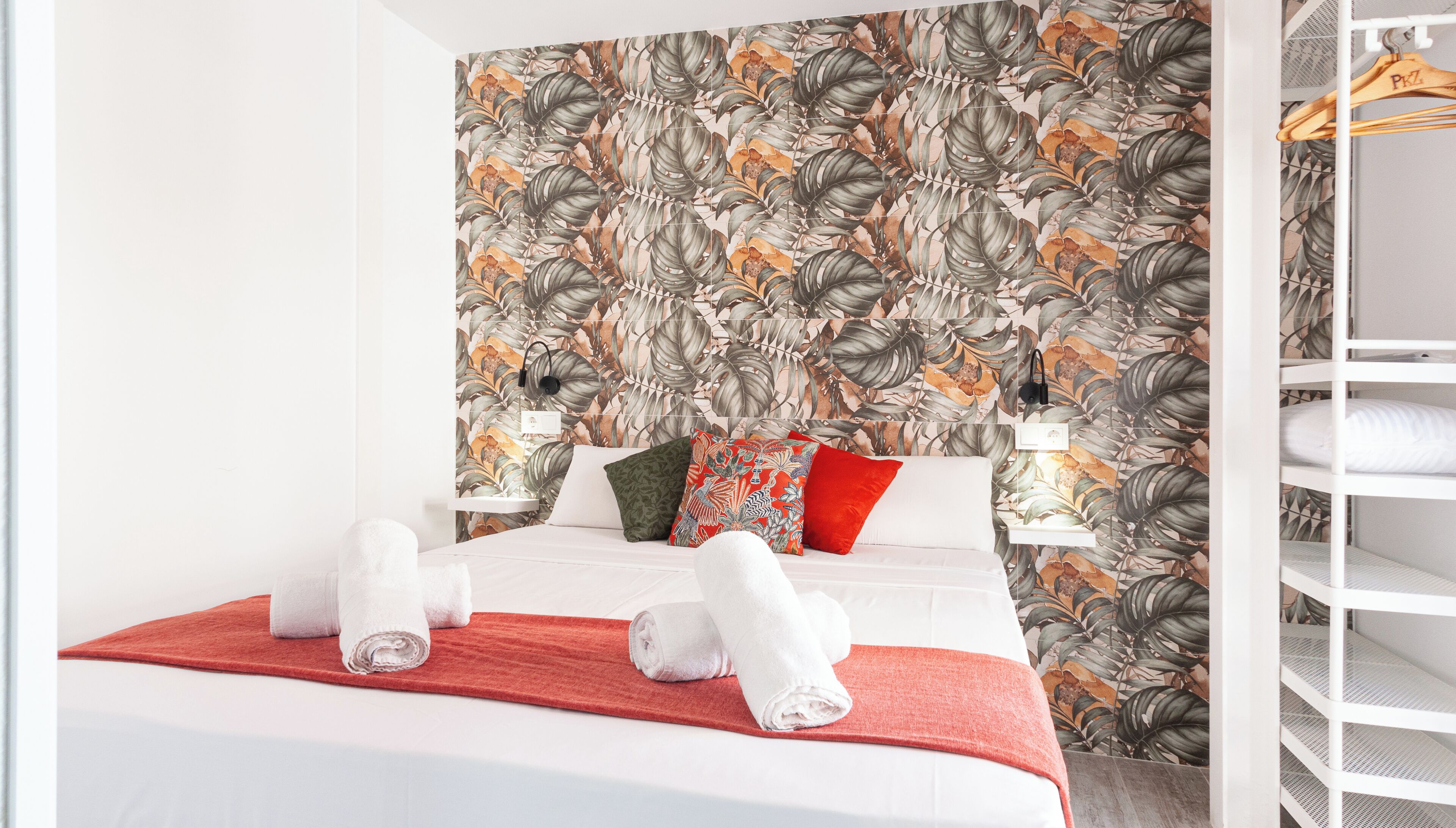 A neatly made bed with white linens and a red throw blanket, a wall with a tropical leaf pattern, and a white shelving unit with hanging clothes.