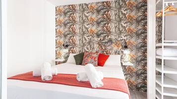 A neatly made bed with white linens and a red throw blanket, a wall with a tropical leaf pattern, and a white shelving unit with hanging clothes.