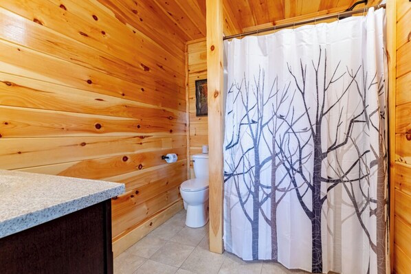 Une salle de bain rustique, aménagée dans une cabane en bois, avec des toilettes, un lavabo et un rideau de douche orné d’un motif d’arbre.