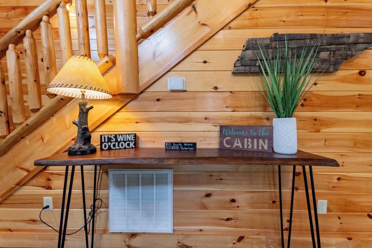 L’intérieur d’un chalet en bois, avec un escalier en bois, une table avec une lampe, une pancarte indiquant « l’heure du vin a sonné », une enseigne de bienvenue et une plante en pot.