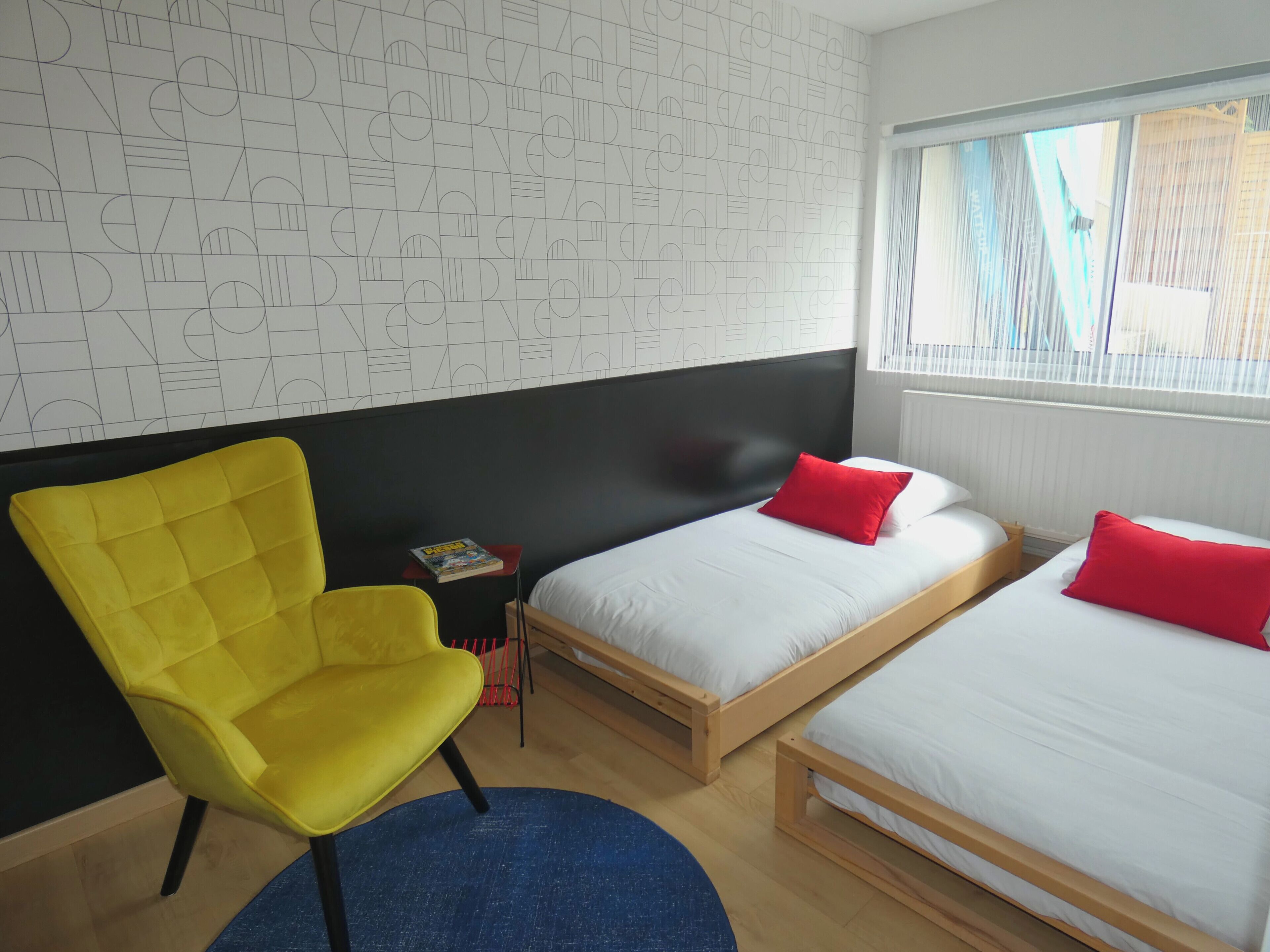 A hotel room with two beds, a yellow chair, a small table with a book, and a patterned wall.