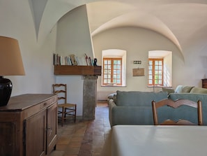 A room with a wooden cabinet, a chair, a sofa, a bookshelf, and two windows with wooden frames.