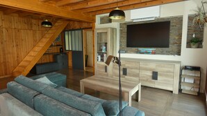Un salón espacioso con un sofá modular grande, techo de madera, televisor de pantalla plana montado en una pared de ladrillo y una escalera que conduce a un nivel superior.