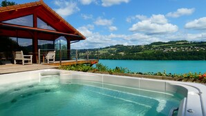Un área de piscina con terraza de madera, sillas de descanso y una estructura de vidrio que da a un lago y colinas.